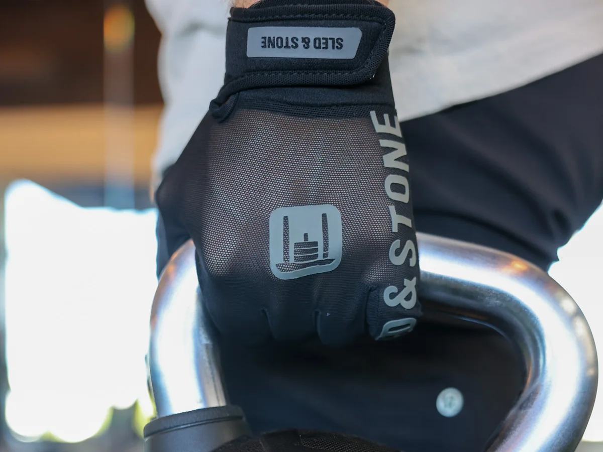 The Stonegrip Glove on a hand gripping a kettlebell handle: wrist tag, sled mark and wordmark down the finger