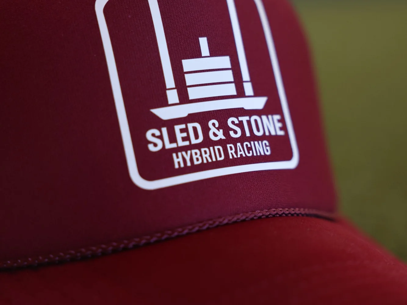 The red Hybrid Racing trucker, laid on turf, with the white Sled & Stone Hybrid Racing patch on the front — view 3